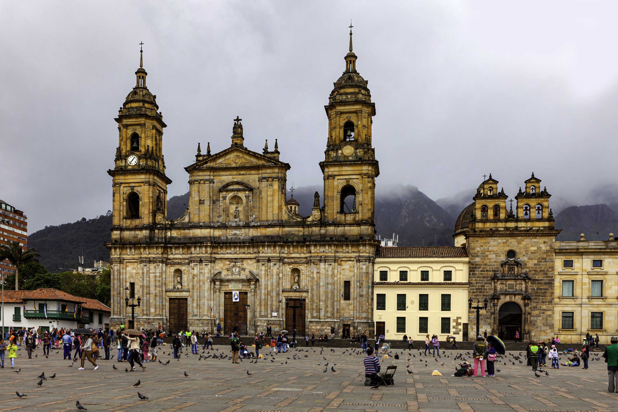 Looking from Plaza Bolivar, in the Andean Capital city of Bogota, in Colombia, South America, to the North Eastern corner of the Square. To the left of the image is the Catedral Primada, the seat of the Roman Catholic Archbishop of Colombia. A few yards the right, is the Archbishop's chapel. A section of his palace can also be seen. The style of architecture is classical colonial Spanish. In the far background in the far background are the Andes Mountains shrouded in rain clouds. There are several local Colombian people in the image; they go about their daily work. The altitude at street level is 8500 feet above mean sea level. Image shot in the afternoon sunlight, on an overcast day; horizontal format. Copy space. Note to Inspector: There are many people in the image, but none of them is more promiment than the other.